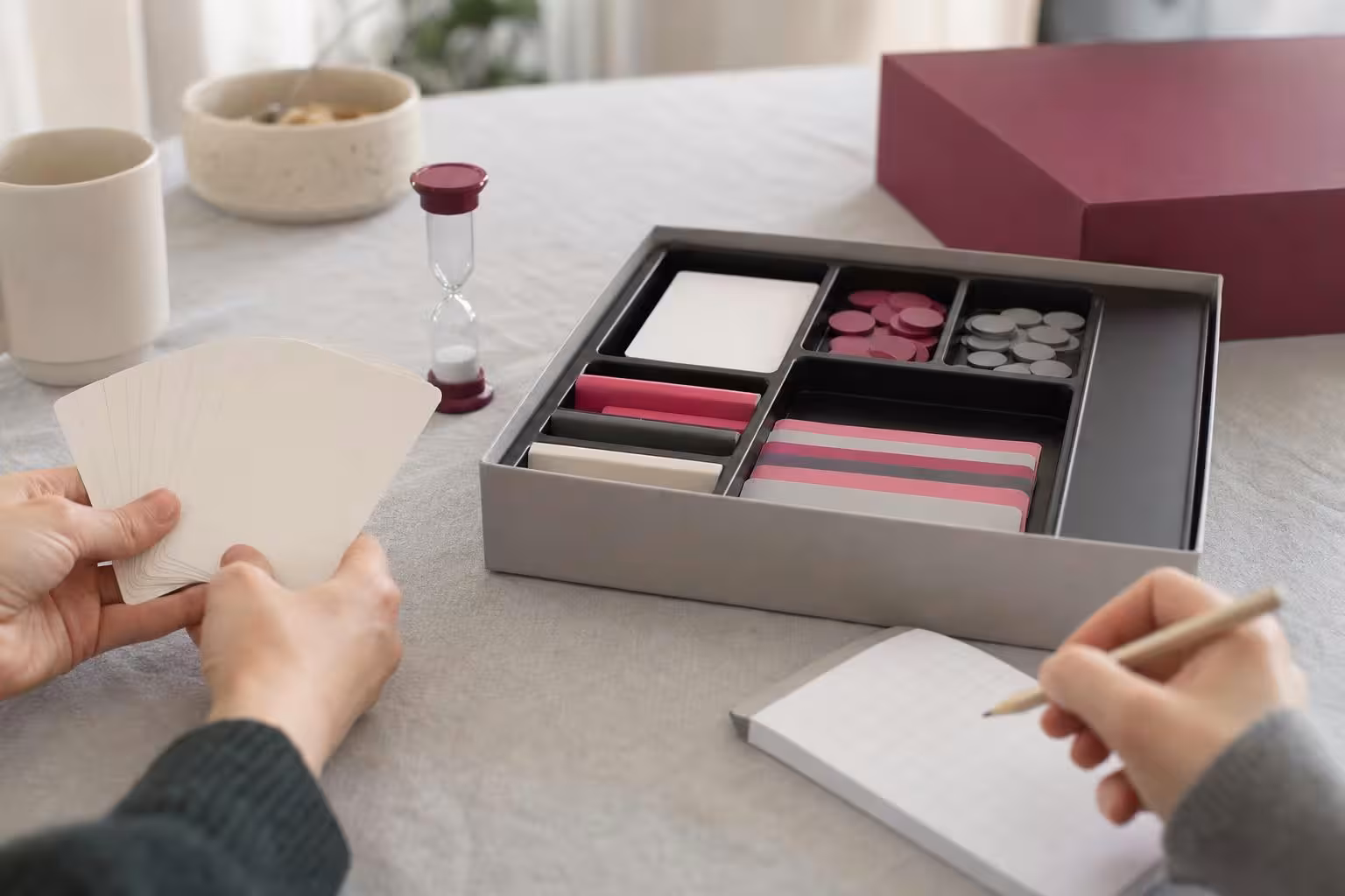 Geöffnete Spieleschachtel mit sortiertem Material, Karten in der Hand und ein Wertungsblock (mit AI erstelltes Bild)